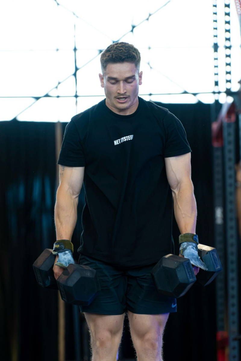 bodybuilder met twee gewichten in de handen en een zwarte t-shirt met het witte logo van Het Systeem