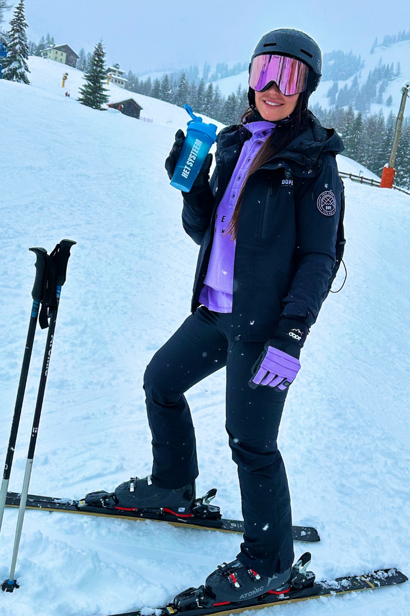 gelukkige jonge vrouw die in de sneeuw op skilatten een shakebeker van het systeem vastheeft