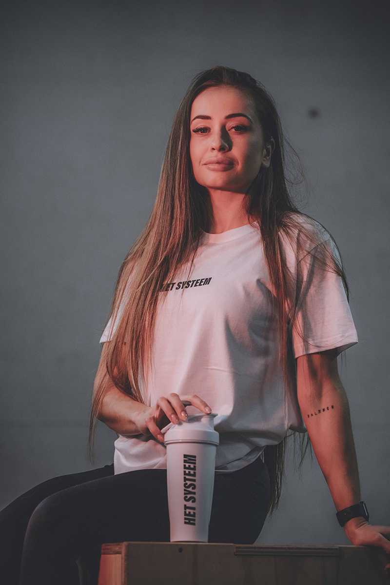 een vrouwelijke personal coach aan het poseren met een shakebeker en t-shirt van het systeen als ambassadeur